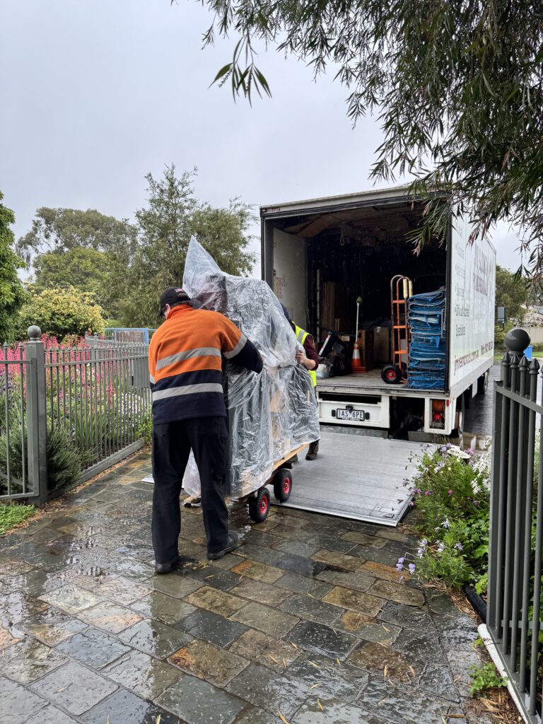 JM & SONS Piano Movers moving a rain-covered grand piano off a hydraulic truck tailgate onto wet paving